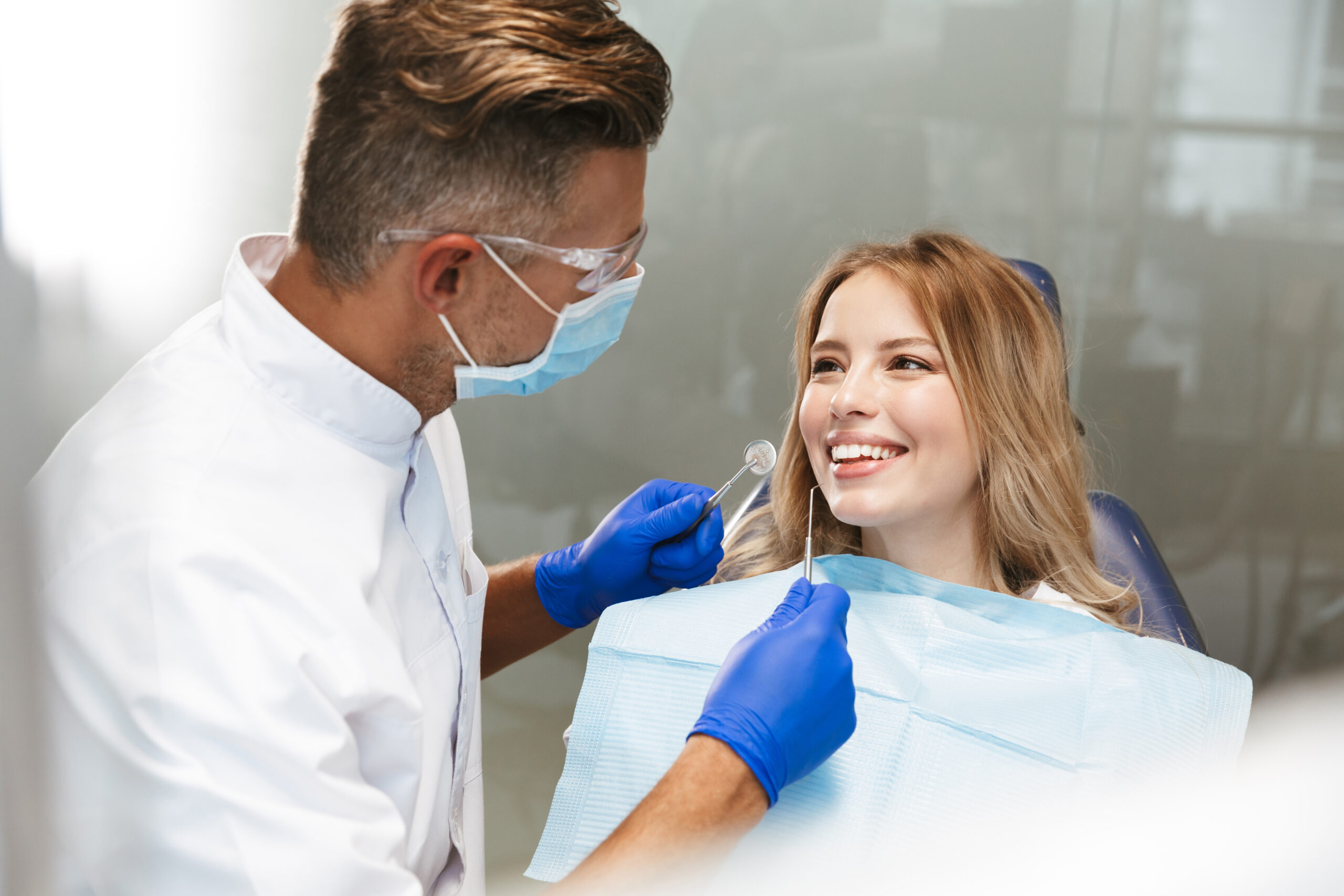 Image of blond young woman sitting in dental chair at medical center while professional doctor fixing her teeth